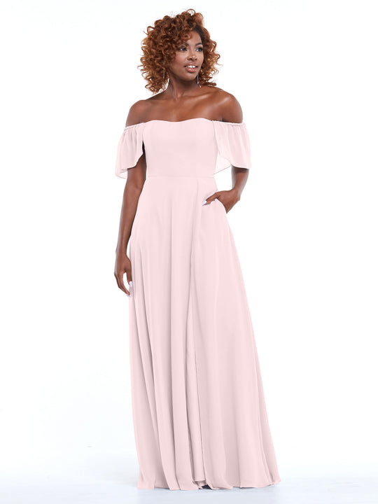 Model wears Avery Austin 80168: a light pink chiffon off-the-shoulder A-line dress with pockets and short ruffled sleeves, posing against a white background with one hand in her pocket and a slight smile.
