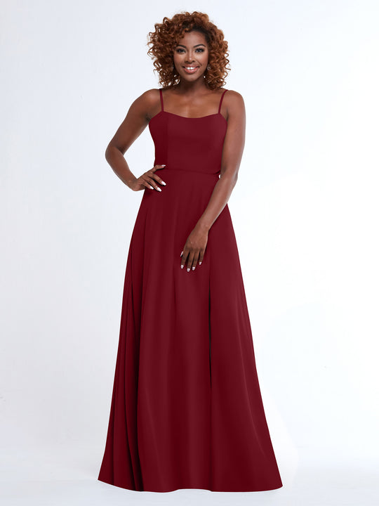 A woman with curly hair smiles in the Avery Austin 80170, a flowy floor-length burgundy A-line dress with spaghetti straps, standing against a plain white background.