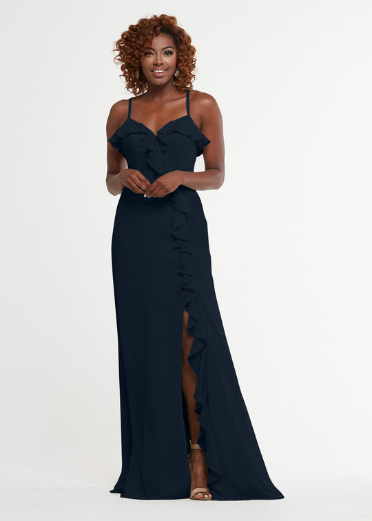 A woman with curly hair smiles as she poses in Avery Austin's 80174—a navy blue, floor-length A-line dress with thin straps, ruffle details, and a side slit—against a plain white background.