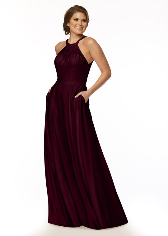 A woman stands against a white background, wearing Avery Austin's maroon A-Line halter neck dress (83002) with a flowy, floor-length skirt and pockets. She smiles slightly, her hair styled in an elegant updo.