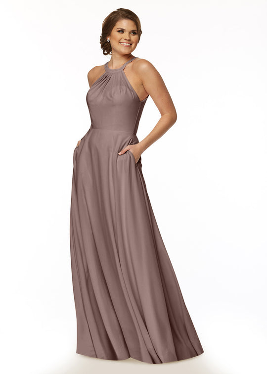 A woman smiles with her hair in a low updo, wearing Avery Austin’s sleeveless mauve A-line dress (style 83002) with a halter neckline and pockets, standing against a white background.