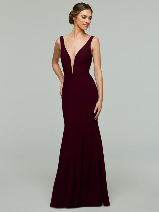 A woman wearing Avery Austin’s 83010, a sleeveless deep burgundy mermaid dress with a plunging V-neckline, stands against a plain gray background, smiling softly with her hair in an elegant updo.