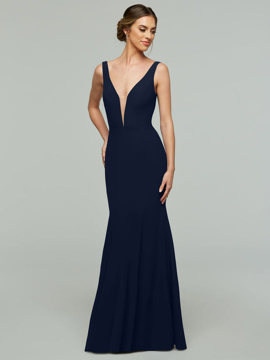 A woman wears the Avery Austin TH-83010, a sleeveless midnight mermaid dress with a plunging V-neckline, standing against a plain light gray background and smiling slightly with one hand on her hip.