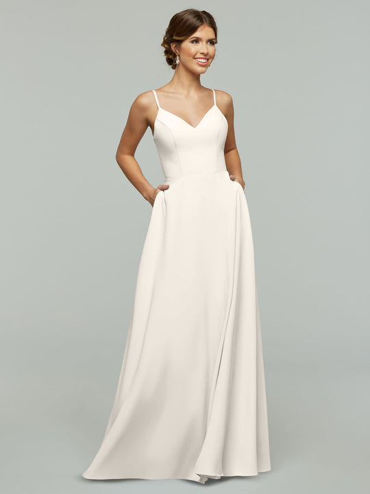 A woman in Avery Austin’s 83012 wears a sleeveless, floor-length white A-line dress with spaghetti straps and pockets, standing against a plain light gray background, smiling and looking slightly to the side.