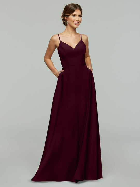 A woman models Avery Austin’s 83012, a sleeveless, floor-length burgundy A-line dress with spaghetti straps and pockets, standing against a light gray background and smiling slightly while looking to the side.