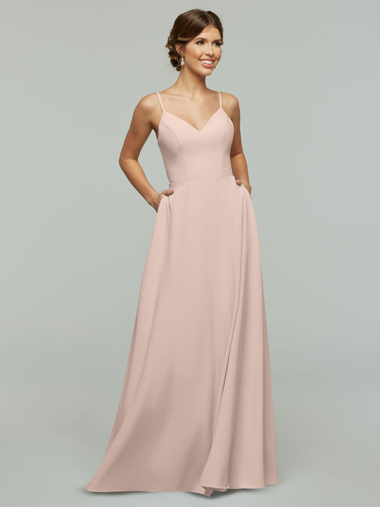 A woman smiles with her hands in the pockets of the Avery Austin 83012, a rose pink floor-length A-line dress with spaghetti straps, standing against a plain light gray background.