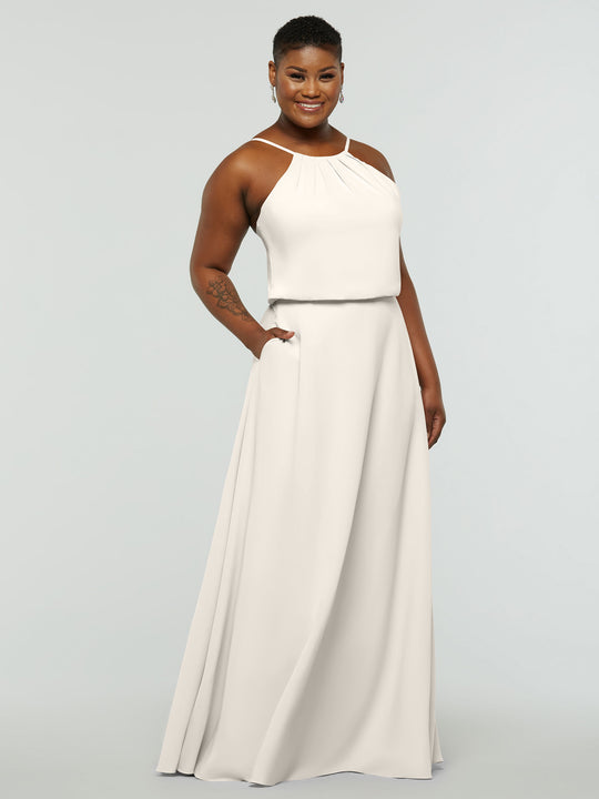 A person smiles in Avery Austin’s 83015—a sleeveless, floor-length cream A-line dress with a high neckline, pleated details, and pockets. They have short hair, earrings, and stand against a plain light gray background.