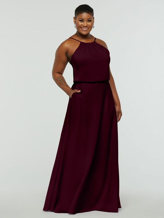 A person smiles in Avery Austin's 83015: a sleeveless, floor-length dark burgundy A-Line dress with a high neckline and side pockets. They have short hair, visible earrings, and stand against a plain light gray background.