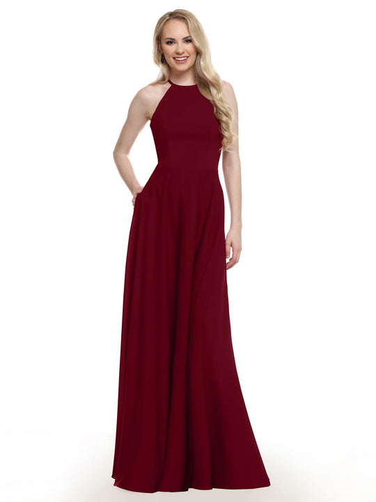 A woman with long blonde hair models the Avery Austin TH-83022, a sleeveless, floor-length dark red A-Line gown. She smiles with one hand in her pocket against a plain white background.