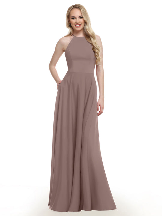 A woman with long, wavy blonde hair smiles in the Avery Austin 83022 gown, showcasing its mauve color, A-line skirt with pockets, and fitted bodice. She poses with one hand in a pocket against a plain white background.