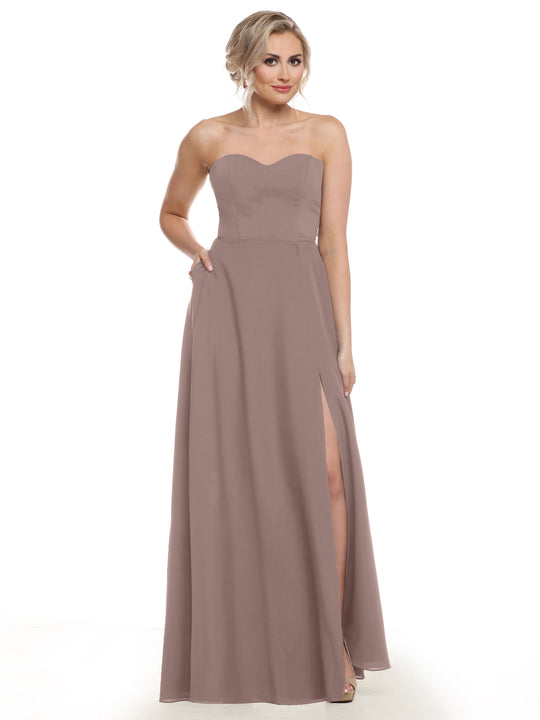 A woman models Avery Austin’s 83025: a strapless chiffon bridesmaid dress with a sweetheart neckline, thigh-high slit, and pockets, standing elegantly against a white background.