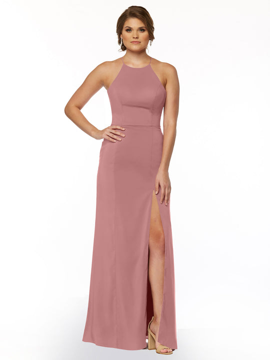 A woman poses against a white background in Avery Austin’s 83026—a rose pink, floor-length sheath dress featuring a halter neck and front slit. She finishes the look with nude heels and one hand on her hip.