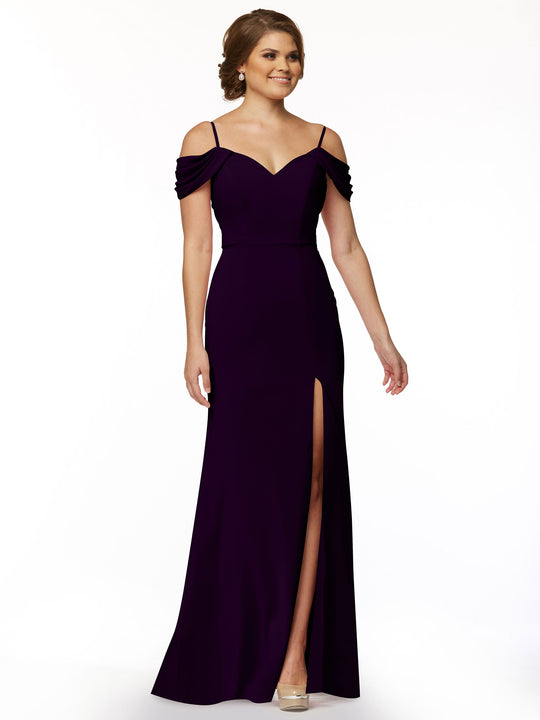 A woman in Avery Austin’s floor-length, fitted off-the-shoulder dress (style 83029) with spaghetti straps and a thigh-high slit poses against a white background. She has short brown hair and beige high heels.