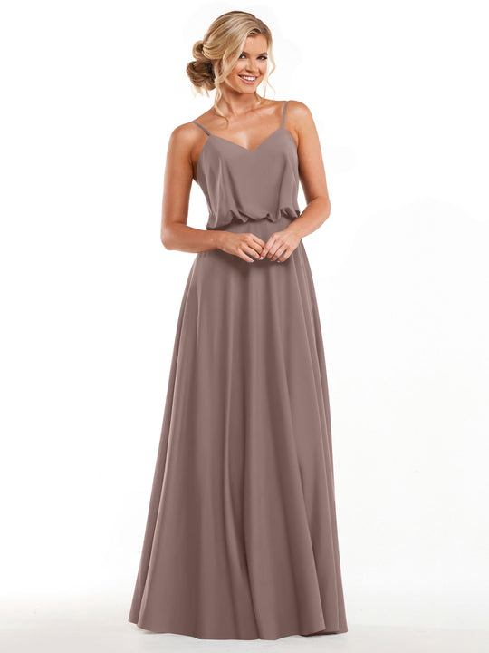 A woman smiles with her hands clasped while modeling Avery Austin’s 83040, a long taupe A-line dress with thin straps and a flowy skirt, against a plain white background.