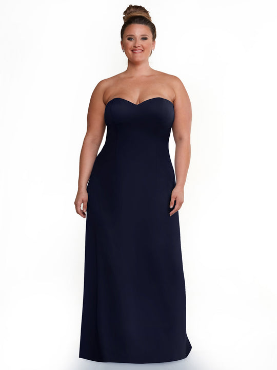 A woman with her hair in a bun smiles while modeling Avery Austin’s 83044, a strapless sweetheart A-line dress in Navy, against a plain white background.