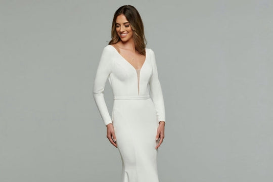 Woman wearing a simple long sleeve white wedding dress with a plunging neckline with a grey background.