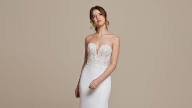 Bride wearing a strapless white wedding dress standing in front of a solid neutral background.