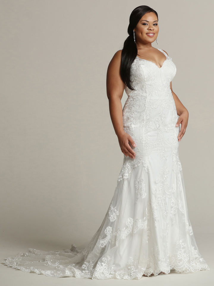 A woman poses in a studio, smiling confidently in the Avery Austin "Evelyn - Tried On Sample," an ivory lace wedding dress featuring a fitted bodice, flowing train, and elegant sleeveless design. Her long dark hair is styled to one side.