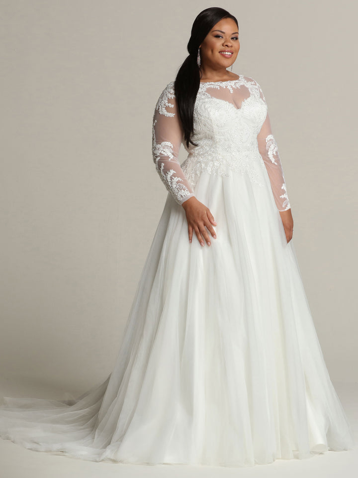 A woman stands smiling against a neutral background, wearing the Avery Austin "Victoria" wedding dress in ivory, featuring long lace and sheer sleeves.