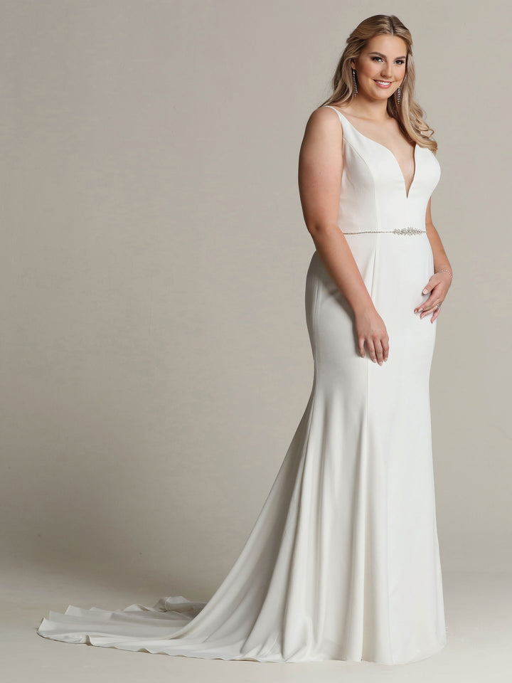 A woman smiles softly in front of a neutral background, wearing the Avery Austin "Aria"—a sleeveless, fit-and-flare wedding dress with a deep V-neckline, beaded waist detail, and long train.