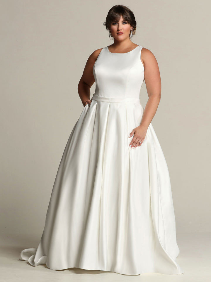 An elegant woman stands against a plain light background, wearing the Avery Austin TH-Penelope—a fitted ivory satin A-line wedding dress with a full pleated skirt and lace back.