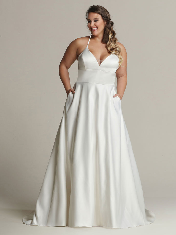A woman smiles against a plain light background wearing Riley - Tried On Sample by Avery Austin, an ivory satin A-line wedding dress with a sleeveless halter neckline and pockets.
