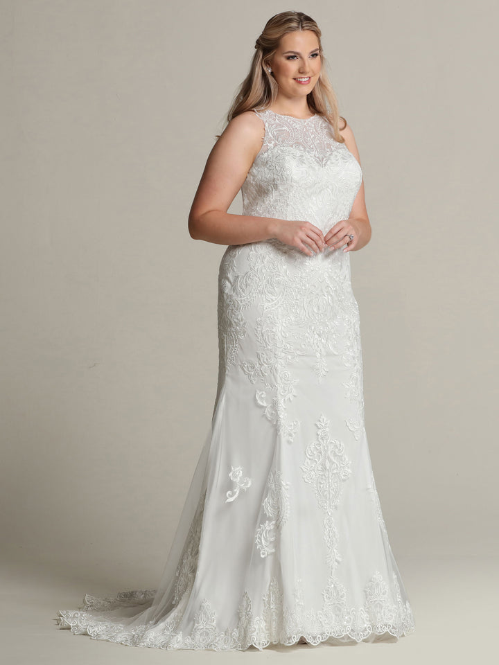 A woman in the Avery Austin TH-Zoey wedding dress, featuring a sleeveless floor-length tulle and lace design with a sheer neckline in Ivory/Ivory, stands against a plain light background, smiling gently with her hands clasped.