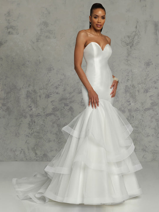 A woman wears the Avery Austin "Natalie" wedding dress—strapless mikado mermaid with a sweetheart neckline and tiered tulle skirt—paired with earrings and a bracelet, standing against a textured gray background, hair styled back.