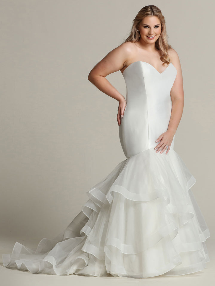 A woman wearing the Avery Austin TH-Natalie ivory mermaid wedding dress, featuring a strapless sweetheart neckline, fitted bodice, and tiered organza ruffled skirt, poses against a plain backdrop with a smile and one hand on her hip.
