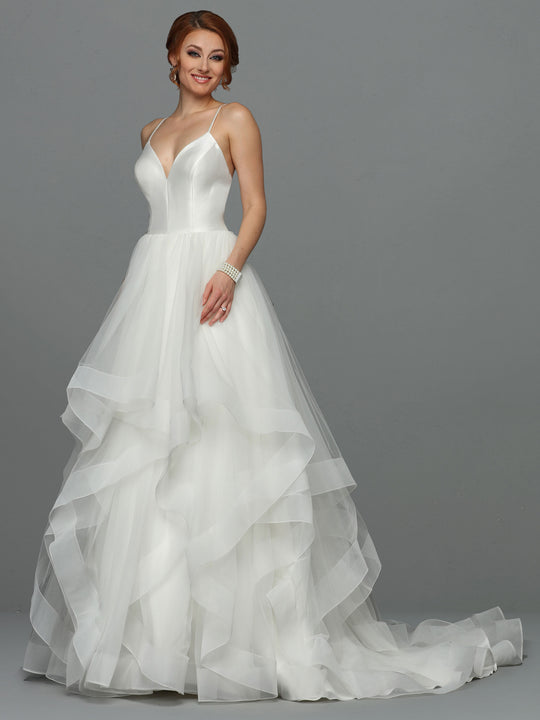A woman smiles against a gray background in the Leah - Tried On Sample by Avery Austin, an ivory wedding dress featuring spaghetti straps, a fitted satin bodice, and a layered tulle skirt. She accessorizes with earrings, a bracelet, and an updo.