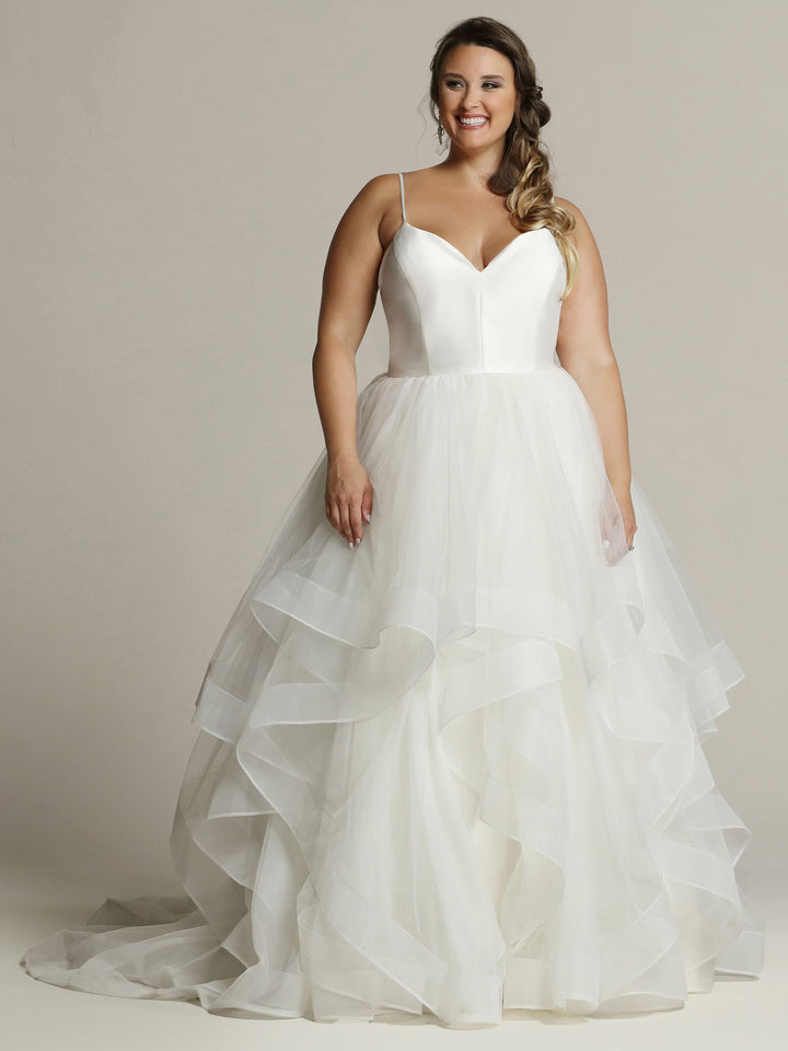 A woman wears the Avery Austin "Leah - Tried On Sample" wedding gown in Ivory, featuring a fitted satin bodice with thin straps and a full layered tulle skirt, smiling against a plain light background.