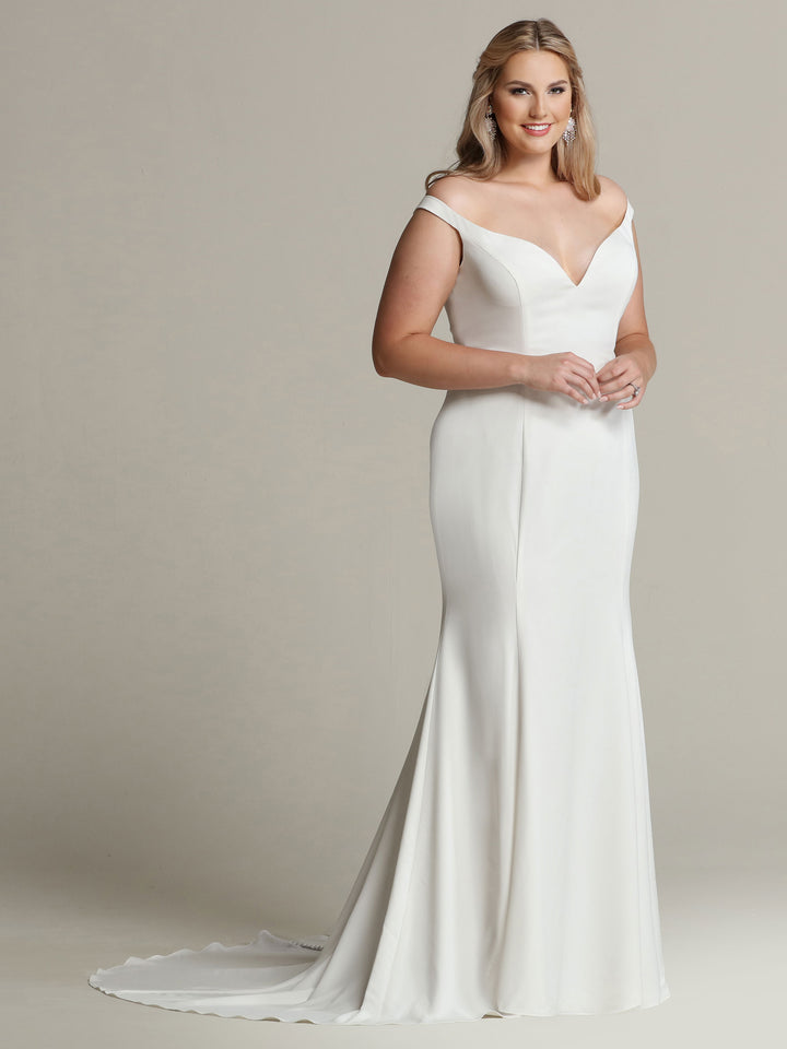 A woman wears the Avery Austin TH-Hazel wedding dress in ivory, featuring an elegant off-the-shoulder crepe design with a fitted bodice and flowing floor-length skirt with a small train. She stands against a plain light background, smiling softly.