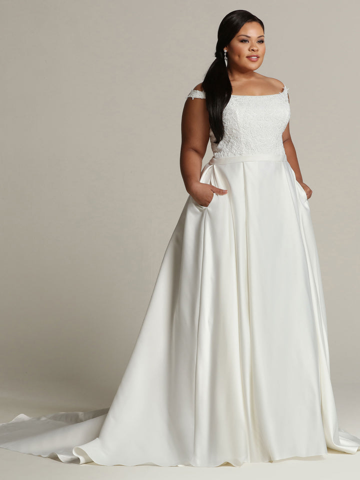 A woman in the ivory "Audrey" gown by Avery Austin, featuring an off-the-shoulder lace bodice and long satin skirt, stands with her hands in her pockets and looks to the side against a plain background.