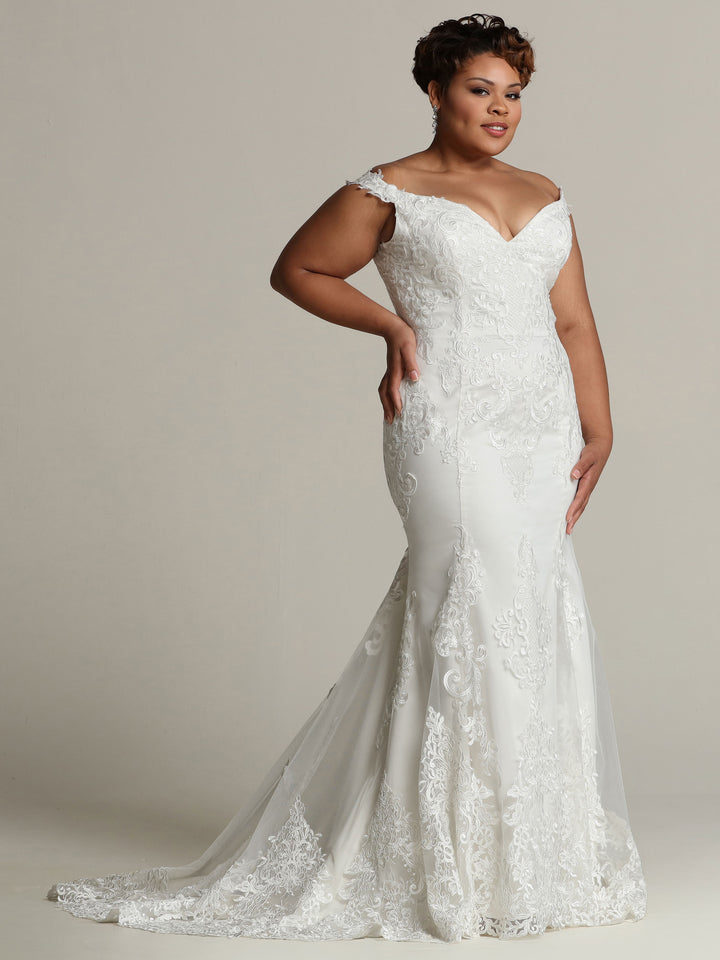 A woman models the Samantha - Tried On Sample by Avery Austin, an ivory/blush off-the-shoulder lace wedding dress with a fitted bodice and dramatic mermaid train, set against a plain light background.