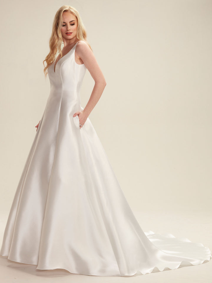 A woman with long blonde hair wears the Avery Austin TH-Maya, a modern sleeveless ivory satin A-line wedding dress with a V-neckline and pockets, standing against a plain light background.