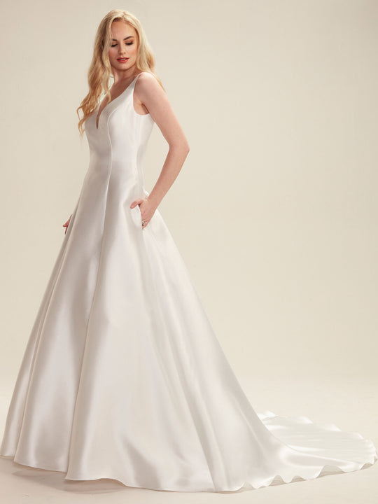 A woman in the Avery Austin "Maya" ivory mikado gown, a sleeveless, floor-length A-line wedding dress with pockets, an open back, long train, and fitted bodice, stands against a plain light background looking slightly down.