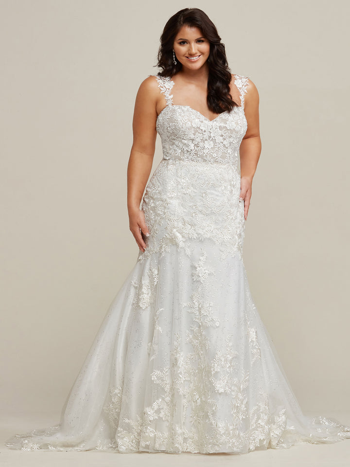 A smiling woman with long, dark hair wears the Avery Austin TH-Nevaeh lace wedding dress in Ivory/Ivory, featuring a fitted bodice and flared skirt, standing against a plain light background.