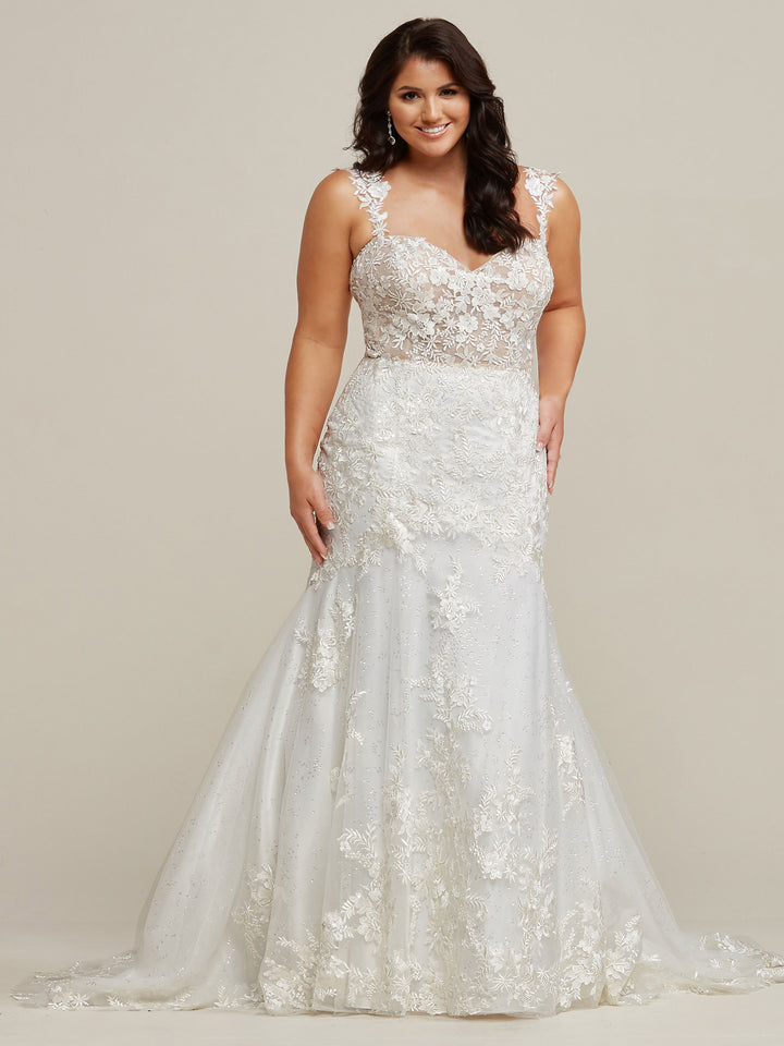 A woman smiles in the Avery Austin Nevaeh wedding dress, an ivory/nude, sleeveless lace gown with a sheer bodice, floral details, and a graceful fit-and-flare silhouette, set against a plain, light background.