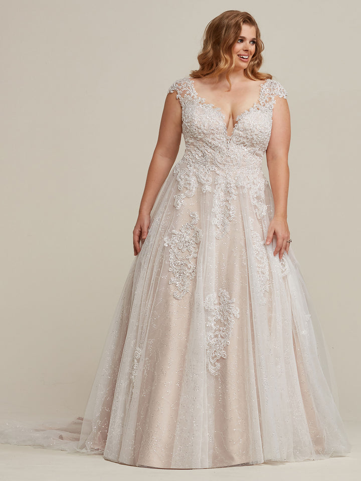 A woman with wavy hair smiles brightly in Avery Austin’s Piper wedding dress, featuring a floor-length A-line silhouette, lace appliqué, deep V-neckline, cap sleeves, and a flowing ivory/champagne tulle skirt against a plain background.