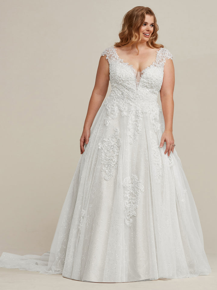 A smiling woman with wavy auburn hair models the Avery Austin TH-Piper wedding dress in Ivory/Ivory, featuring lace appliqué, cap sleeves, a deep V-neckline, and an embellished A-line silhouette against a neutral background.