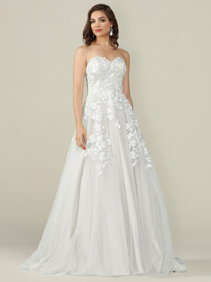 A woman models the Avery Austin TH-Lydia, an ivory strapless floor-length A-line wedding dress with floral lace appliqué and a sweetheart neckline, standing against a plain backdrop.