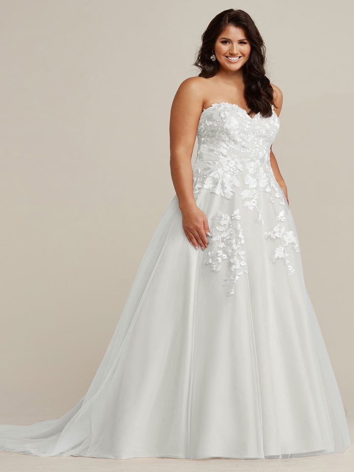 A woman in the Avery Austin TH-Lydia strapless A-line wedding dress in Ivory/Ivory with floral lace appliqué smiles against a neutral background.