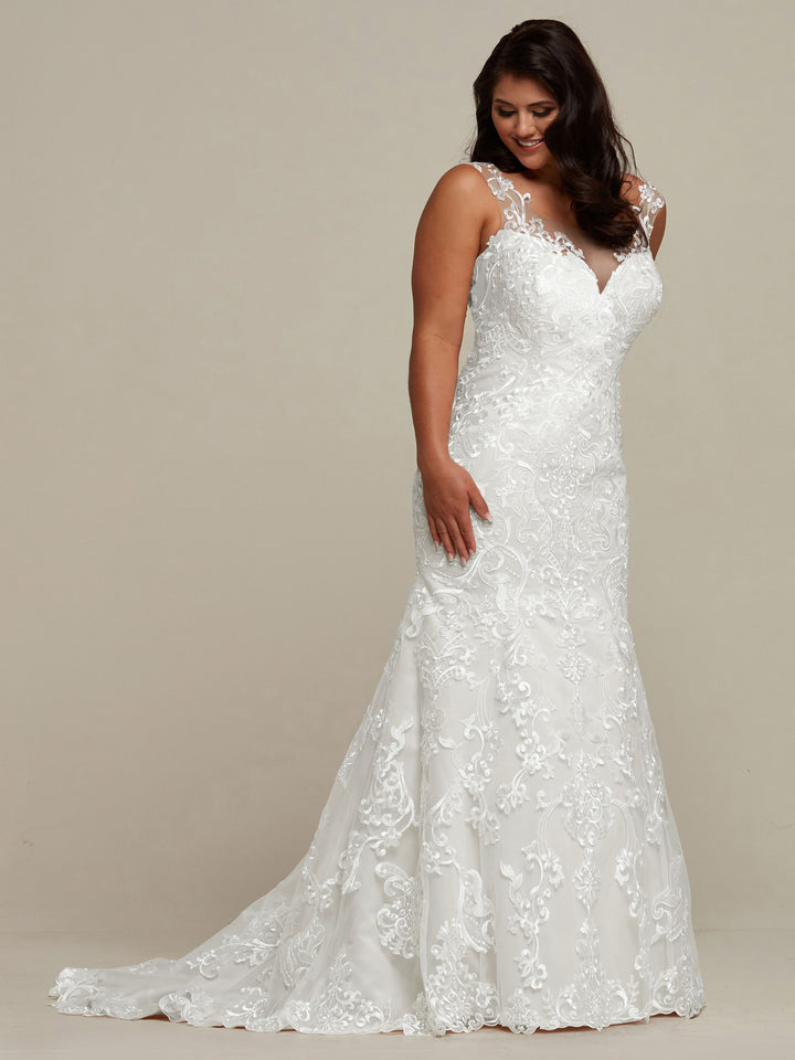 A woman in the Avery Austin TH-Eliana ivory wedding dress—with a fitted lace bodice, sweetheart neckline, and slight train—smiles gently, her hands resting on her gown against a neutral backdrop.