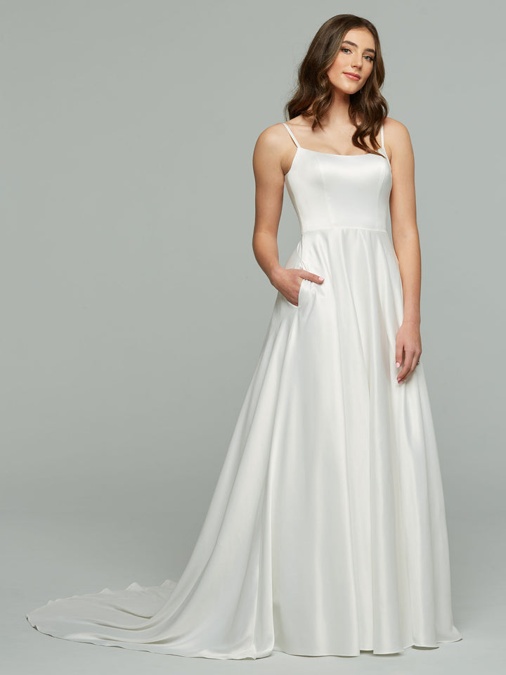 A woman models the averyaustin TH-Giselle, an ivory sleeveless A-line wedding dress with thin straps and pockets, standing against a plain gray background and smiling softly while looking to the side.