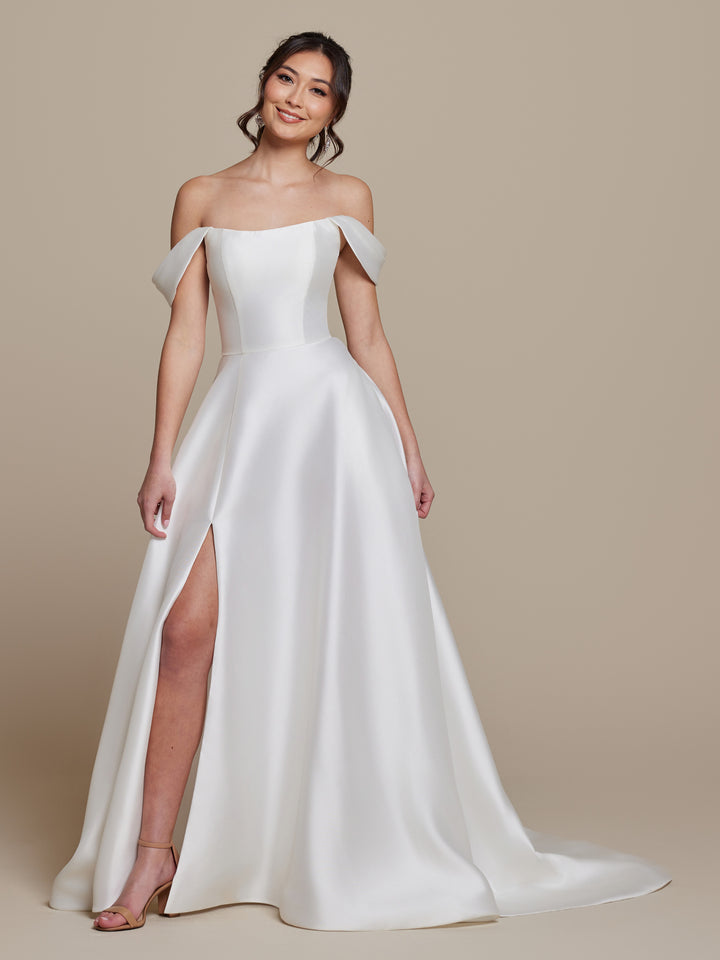 A woman smiles against a beige background, wearing the Poppy ivory off-the-shoulder wedding dress by averyaustin with a thigh-high slit. Her dark hair is styled in loose waves, and she finishes the look with strappy heels.