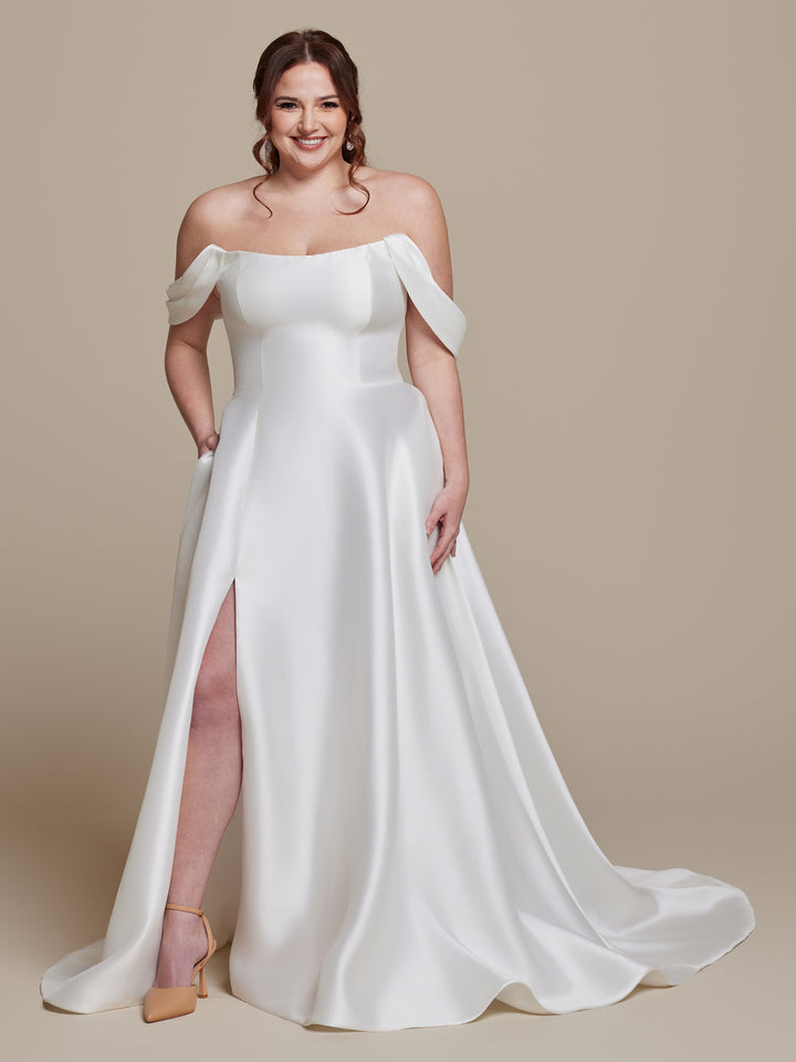 A woman stands smiling with hands in pockets against a beige background, wearing the ivory "Poppy" off-the-shoulder wedding dress by averyaustin, featuring a thigh-high slit, draped sleeves, and modern style. She completes the look with nude heels and loose waves.