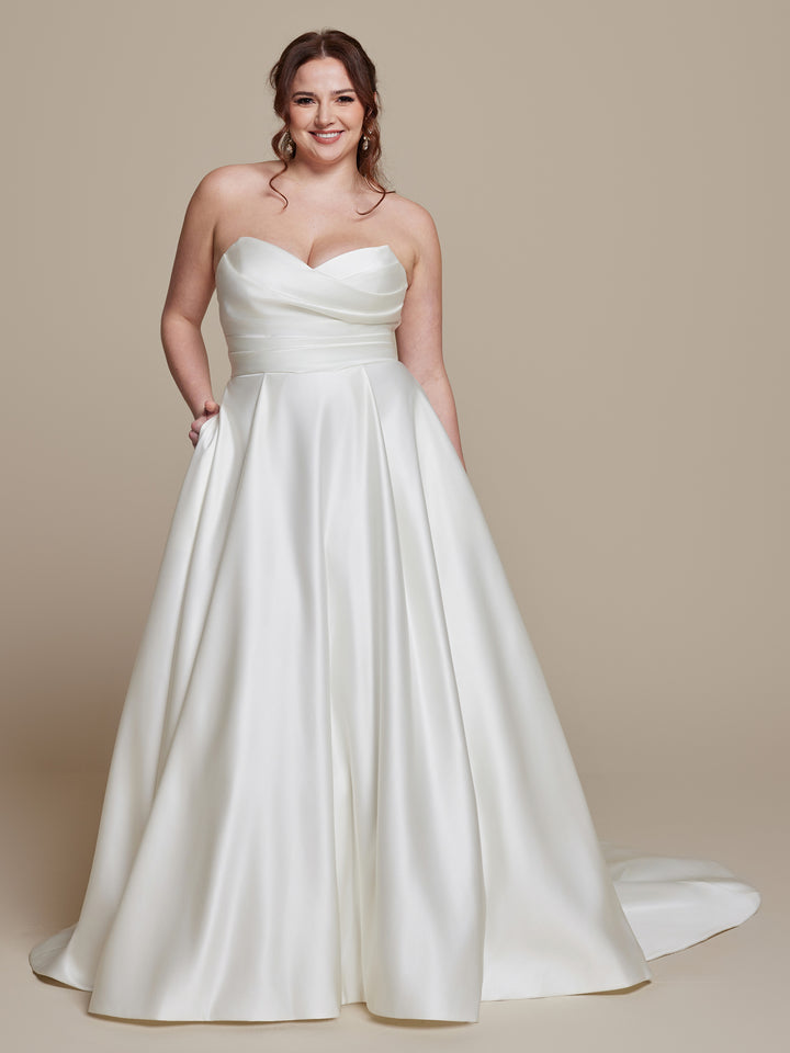 A woman smiles in the "Rosie" by averyaustin, an ivory strapless satin wedding dress featuring a sweetheart neckline, fitted bodice, full floor-length skirt, and side pockets, posing against a plain beige background.