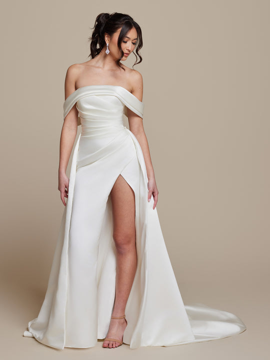 A woman in the ivory Phoebe off-the-shoulder wedding dress by averyaustin, featuring a high slit, draped bodice, and long train, stands against a beige background. She has wavy hair in a ponytail and wears drop earrings and strappy heels.