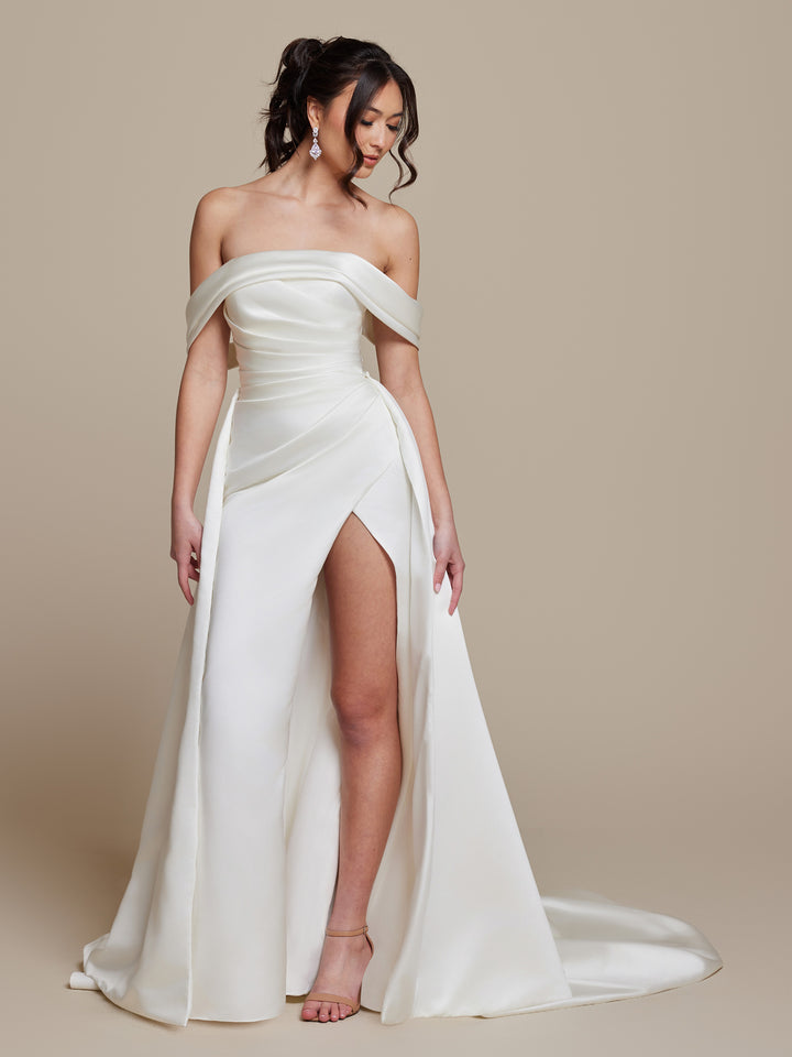 A woman in the ivory Phoebe off-the-shoulder wedding dress by averyaustin, featuring a high slit, draped bodice, and long train, stands against a beige background. She has wavy hair in a ponytail and wears drop earrings and strappy heels.