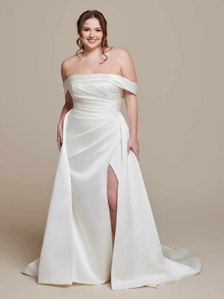 A woman smiles in the ivory Phoebe off-the-shoulder satin wedding dress by averyaustin, featuring a thigh-high slit, against a plain beige background.